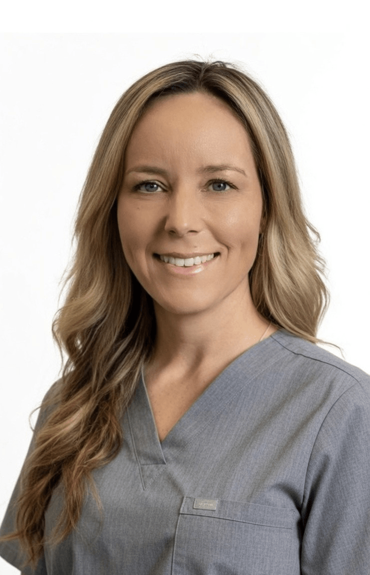 A blonde woman dressed in grey medical scrubs smiles at the camera.
